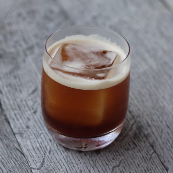 A dark amber cocktail with a large ice cube and white foam on a gray wooden surface.