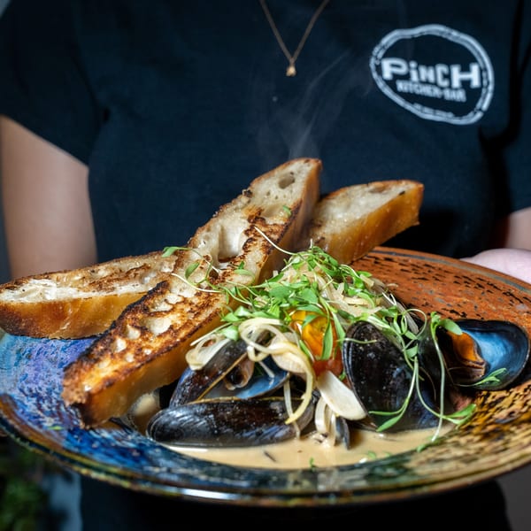 Steaming mussels in broth with grilled bread and microgreens, held by a person in a Pinch Kitchen+Bar shirt.