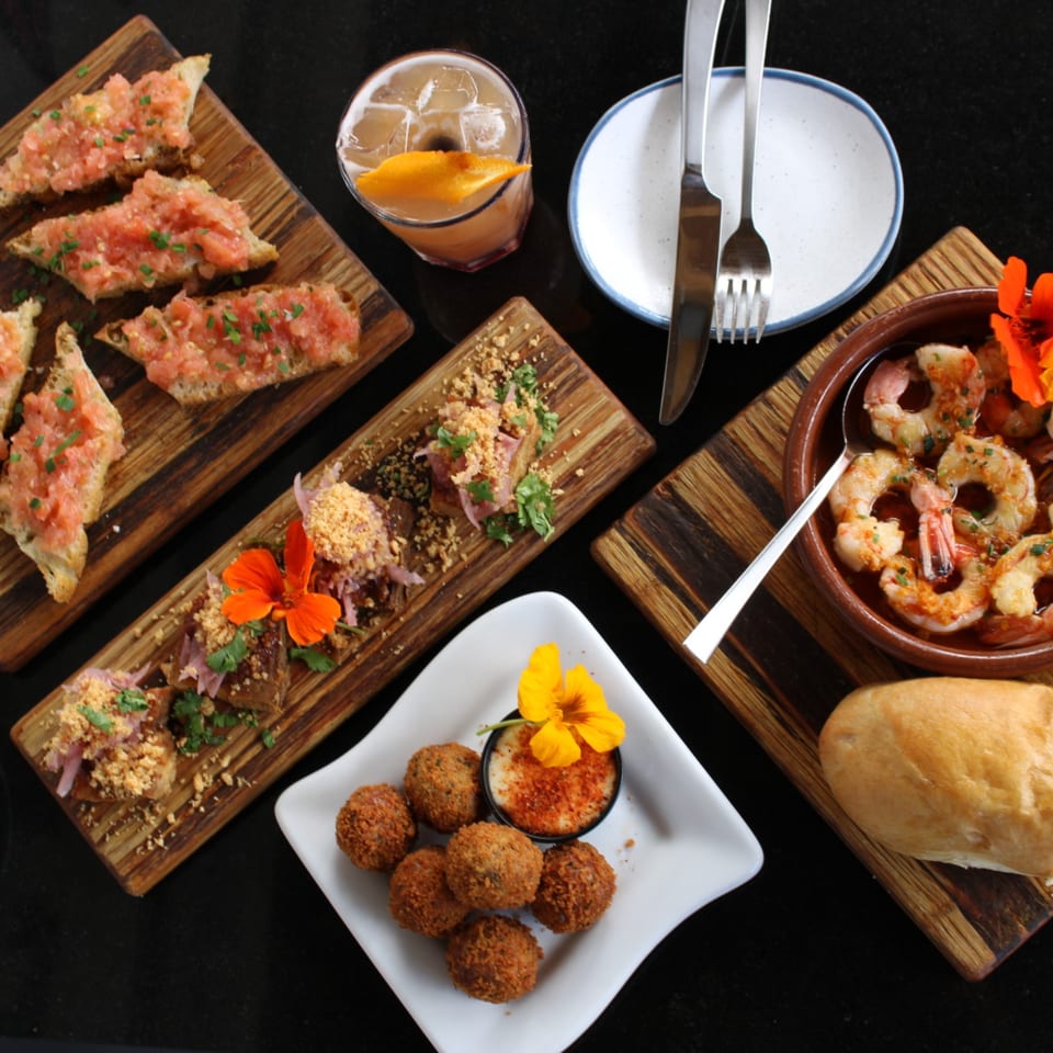An overhead view of Pinch Kitchen+Bar's American tapas, including bruschetta, shrimp, croquettes, and a cocktail.