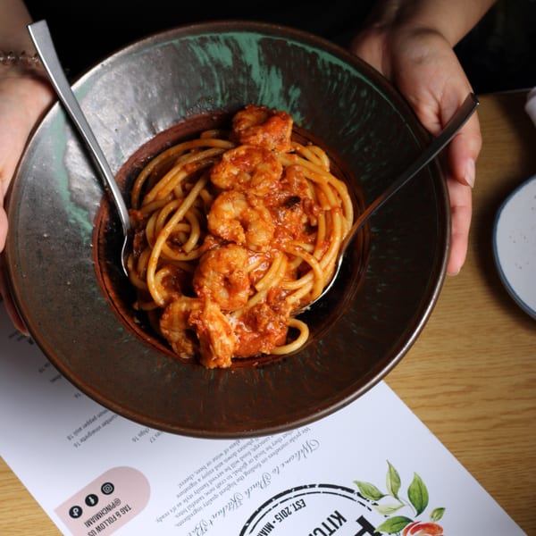 Hands holding a dark bowl of Pinch Kitchen+Bar's shrimp pasta with red sauce and two spoons.
