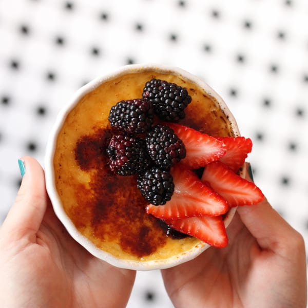 Hands holding a Pinch Kitchen+Bar creme brulee topped with fresh blackberries and sliced strawberries.